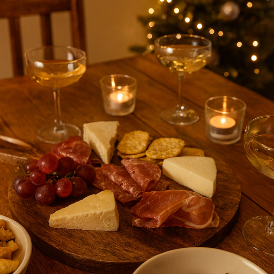 Wooden charcuterie board with meats, cheeses, and grapes on a table with glasses of white wine and candles in the background.