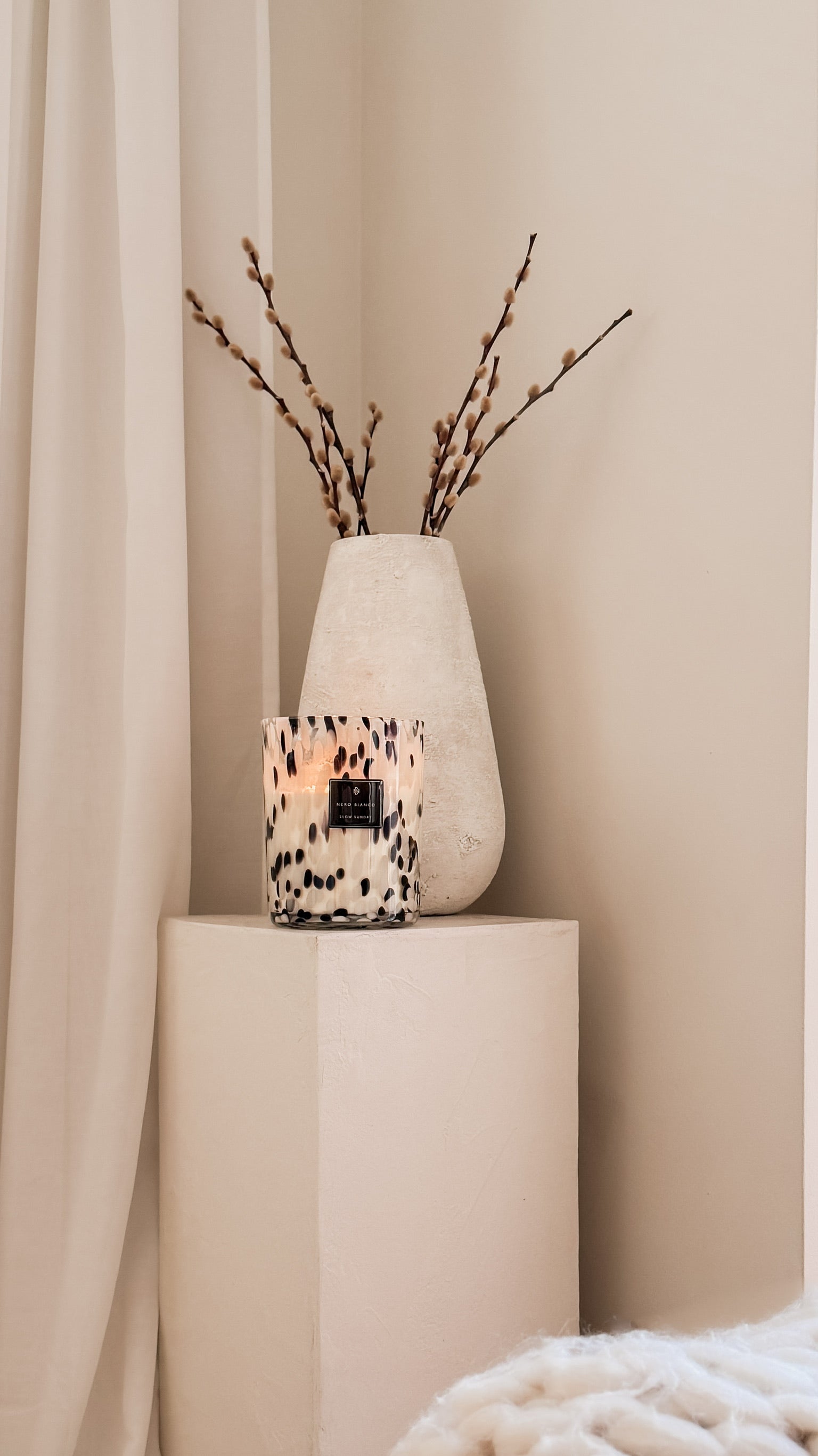 Candle in a decorative vase with branches on a beige pedestal against a beige curtain.