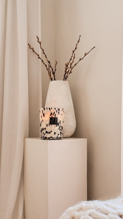 Candle in a decorative vase with branches on a beige pedestal against a beige curtain.