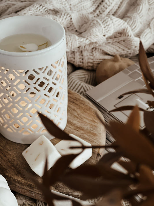 White ceramic wax melt burner with a lit candle on a wooden surface, surrounded by soft lighting and textures.
